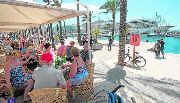 Algunos de los turistas que llegaron ayer en los cruceros 'Ventura' y 'Viking Sky', sentados en una terraza del puerto.Al fondo los dos barcos. /Antonio Gil / AGM